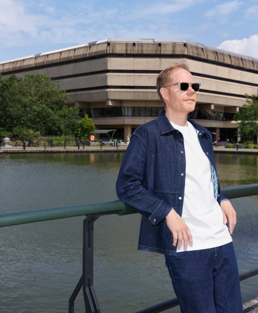 Adult male poses by a canal in full denim outfit with sunglasses, framed by buildings and greenery under a bright, cloudy sky.
