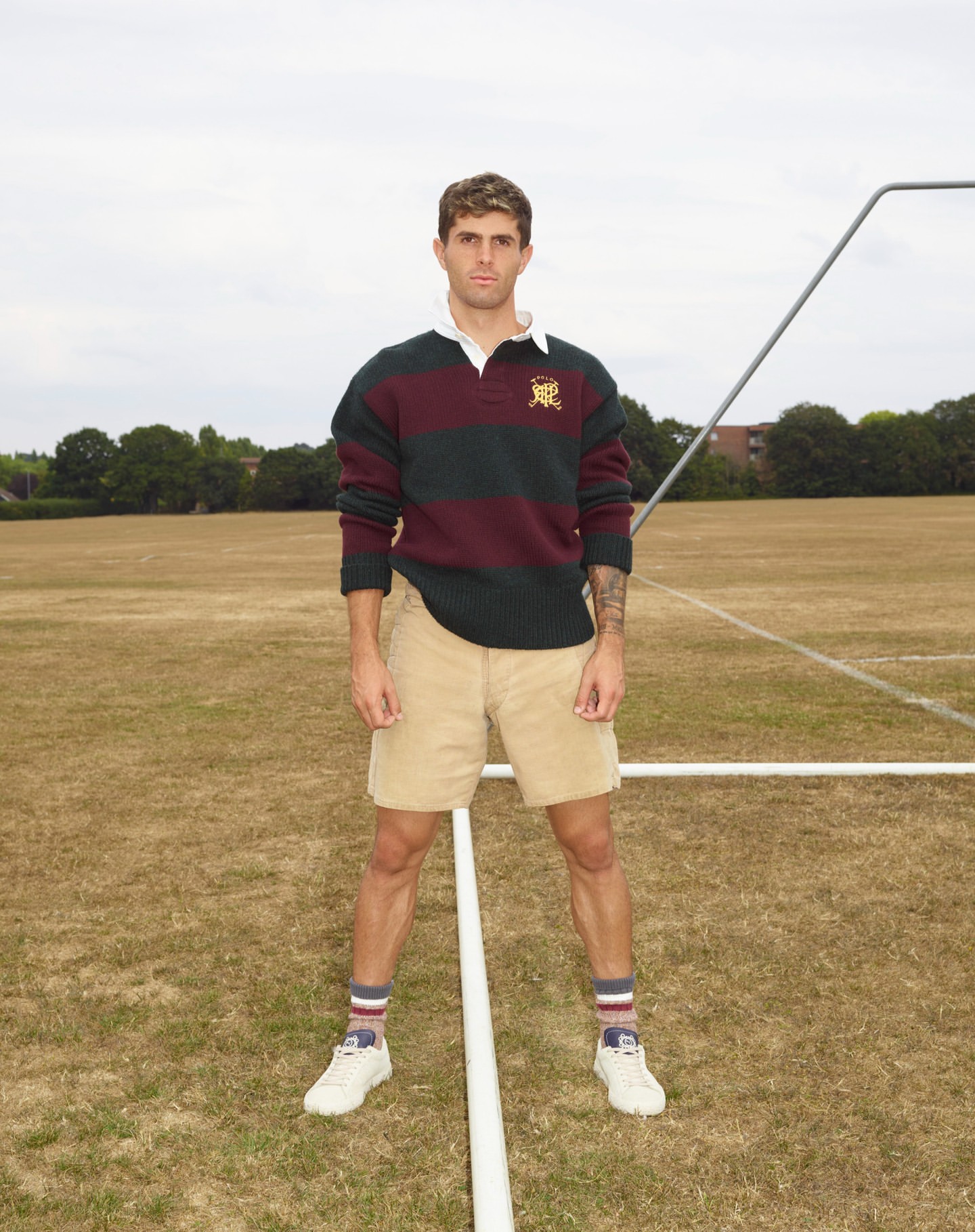 Male model in athletic wear poses on a sports field with white sneakers, striped socks, tan shorts and a striped rugby shirt.
