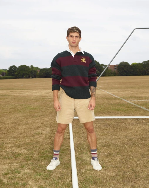 Male model in athletic wear poses on a sports field with white sneakers, striped socks, tan shorts and a striped rugby shirt.