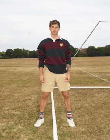 Male model in athletic wear poses on a sports field with white sneakers, striped socks, tan shorts and a striped rugby shirt.