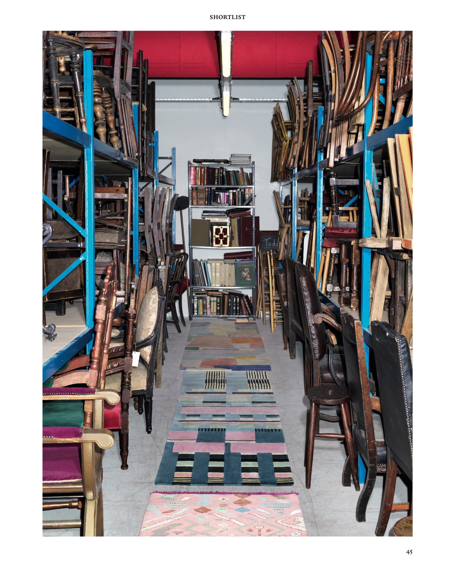 Indoor storage room filled with rows of antique chairs on blue metal shelves, with area rugs on the floor between the aisles.