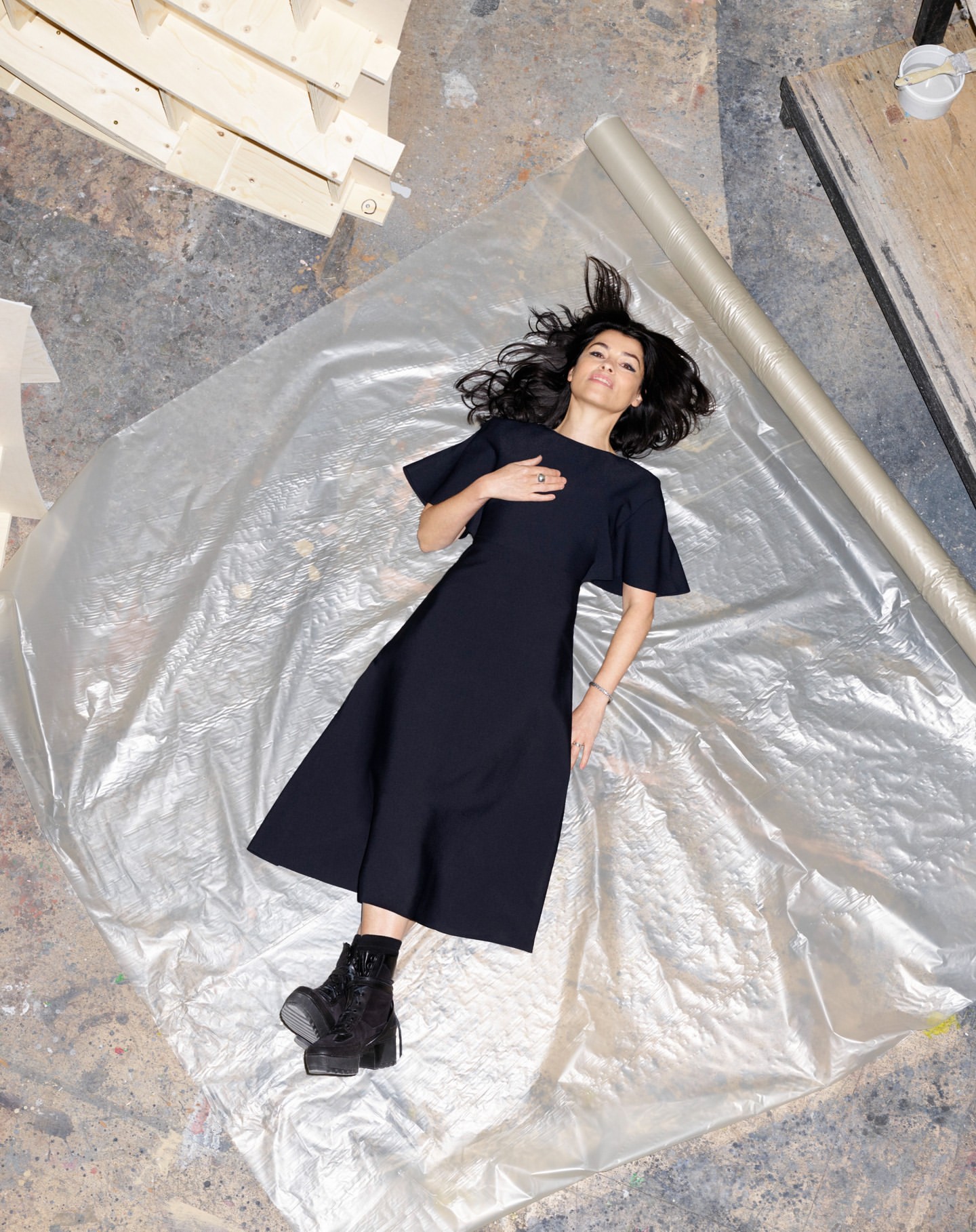 Overhead shot of a woman in a black dress lying on a translucent plastic sheet, wearing black platform boots in a brightly lit room.