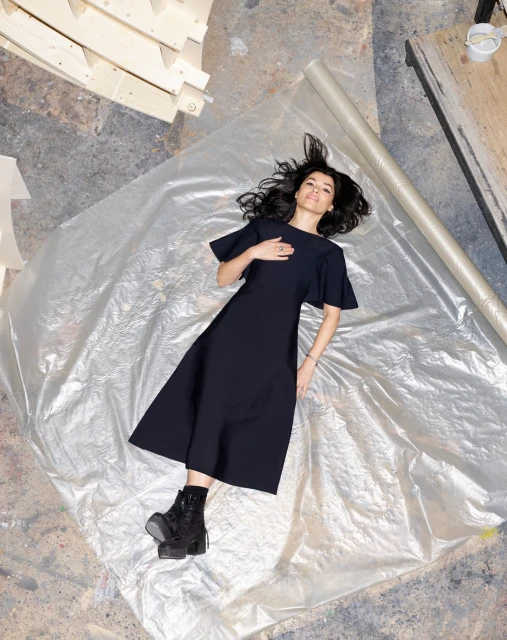 Overhead shot of a woman in a black dress lying on a translucent plastic sheet, wearing black platform boots in a brightly lit room.