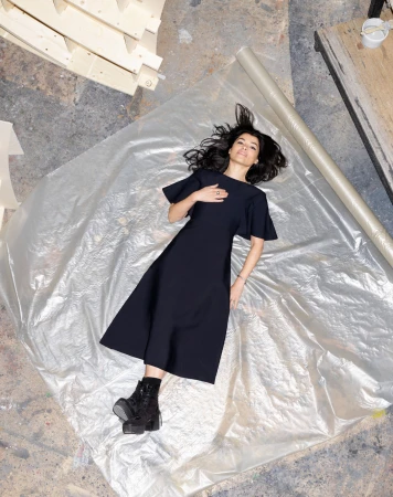 Overhead shot of a woman in a black dress lying on a translucent plastic sheet, wearing black platform boots in a brightly lit room.
