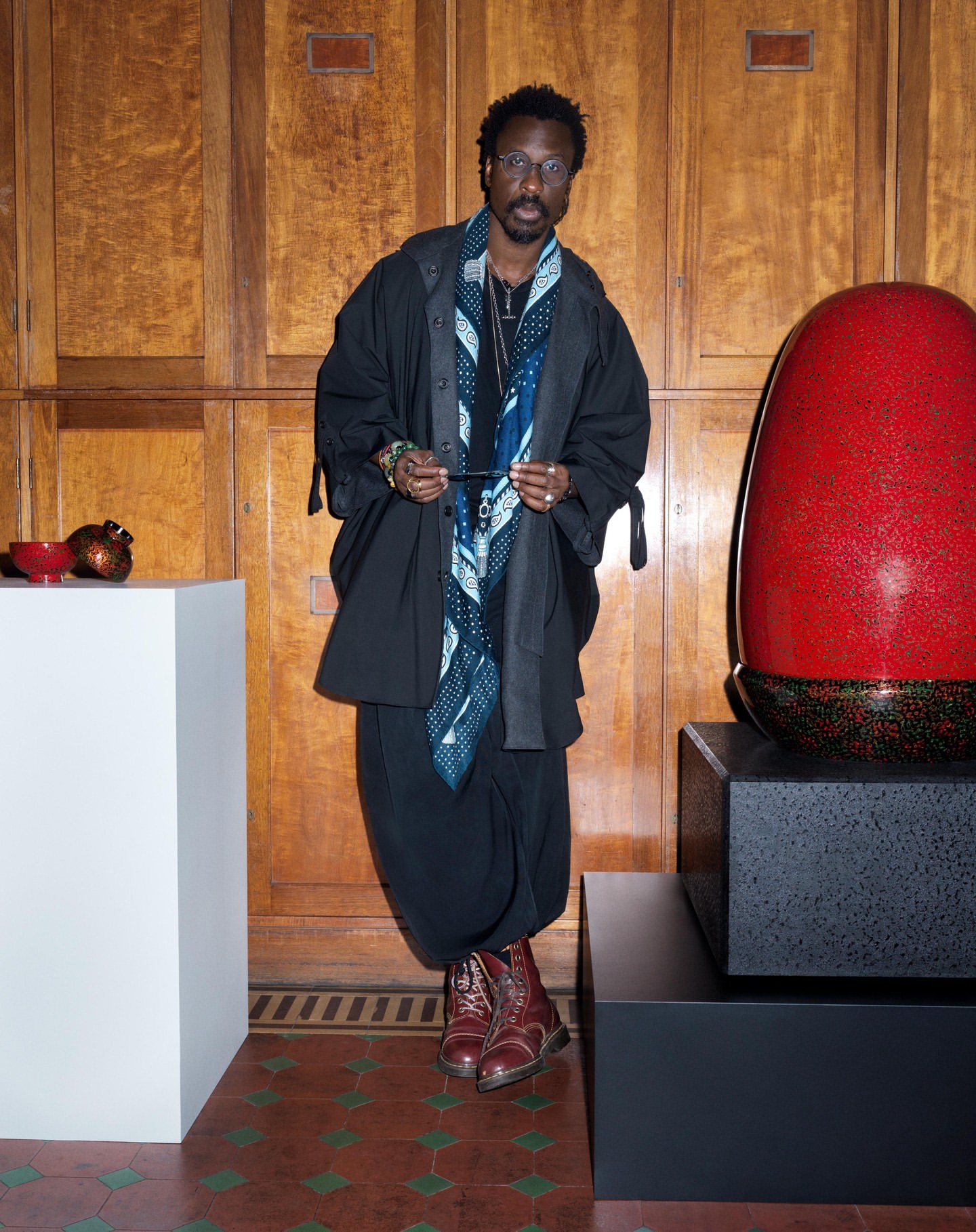 An adult male wearing glasses and a patterned scarf poses in front of a wooden paneled wall and art pieces, set on pedestals.