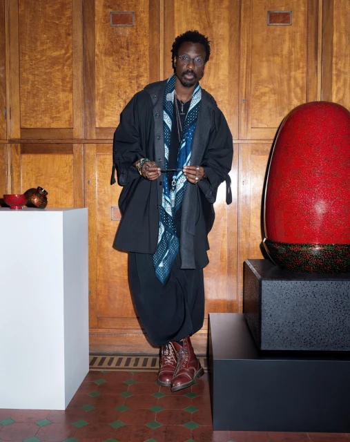 An adult male wearing glasses and a patterned scarf poses in front of a wooden paneled wall and art pieces, set on pedestals.