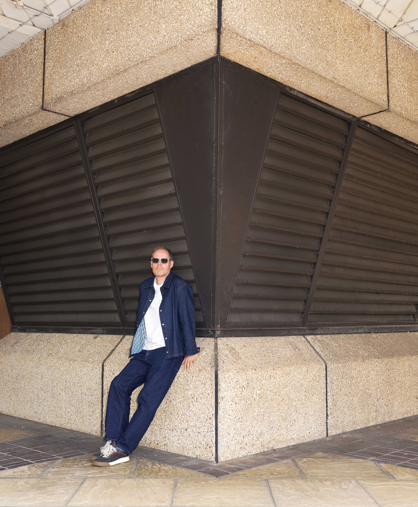 Man wearing a blue denim jacket leaning against a concrete column with dark louvers by photographer Frederike Helwig in London
