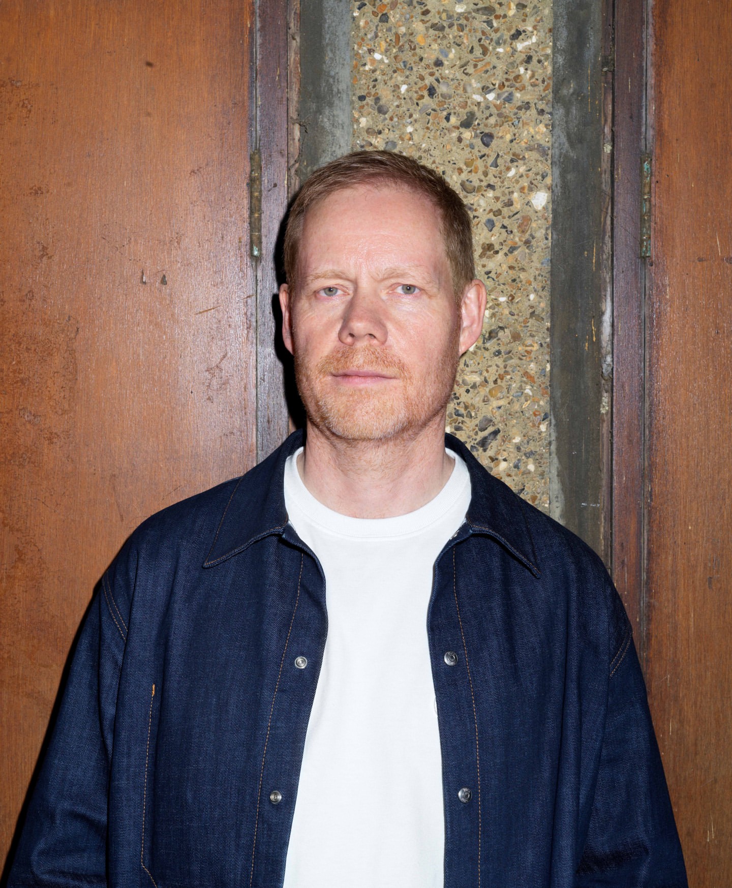 Portrait of a fair-skinned adult male with red hair wearing a white t-shirt and a denim jacket in front of a textured wall.