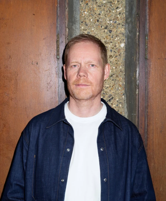 Portrait of a fair-skinned adult male with red hair wearing a white t-shirt and a denim jacket in front of a textured wall.