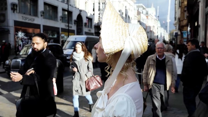Woman in bonnet and white dress stands in urban setting amongst pedestrians and vehicles on city street with shops and buildings.