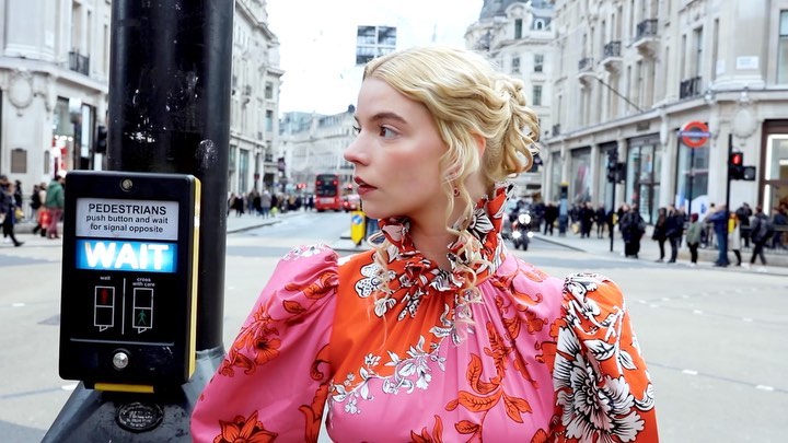 Anya Taylor-Joy is standing on a city street in a floral dress with buildings and people in the background; crossing signal visible.
