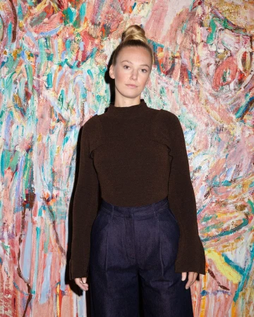 Portrait of a blonde woman wearing a dark brown mock-neck top and dark trousers against an expressive abstract painted background.