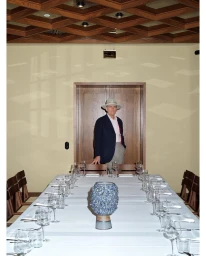 An elderly man in a hat and navy blazer standing at the head of a long dining table set with glassware.