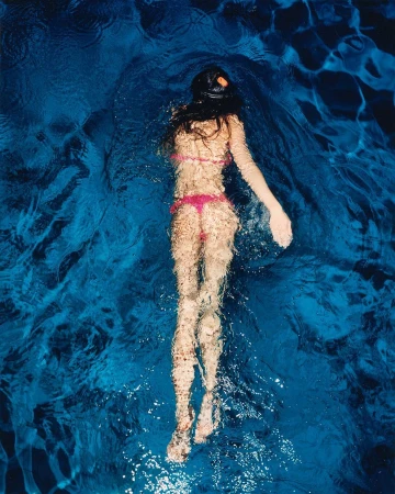 Woman wearing a pink bikini swimming underwater in a deep blue pool captured from overhead by Frederike Helwig