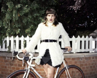 Woman in white fluffy coat with black belt standing with a white bicycle in front of a white picket fence.