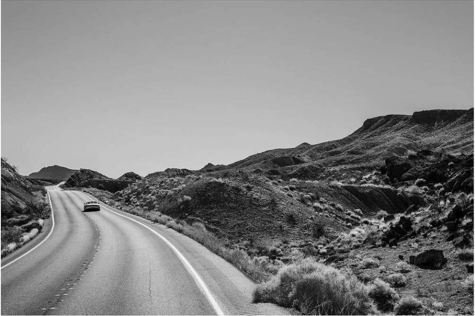 A black and white landscape photograph shows a car on an open road leading through a hilly desert landscape under a clear sky.