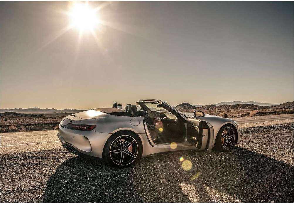Silver convertible parked on a roadside with distant mountains under a sunny sky. The car door is open, inviting exploration.