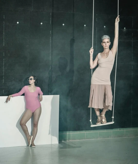 Two women pose in a photo studio: one in a pink leotard leans on a white block, the other in beige attire sits on a swing.