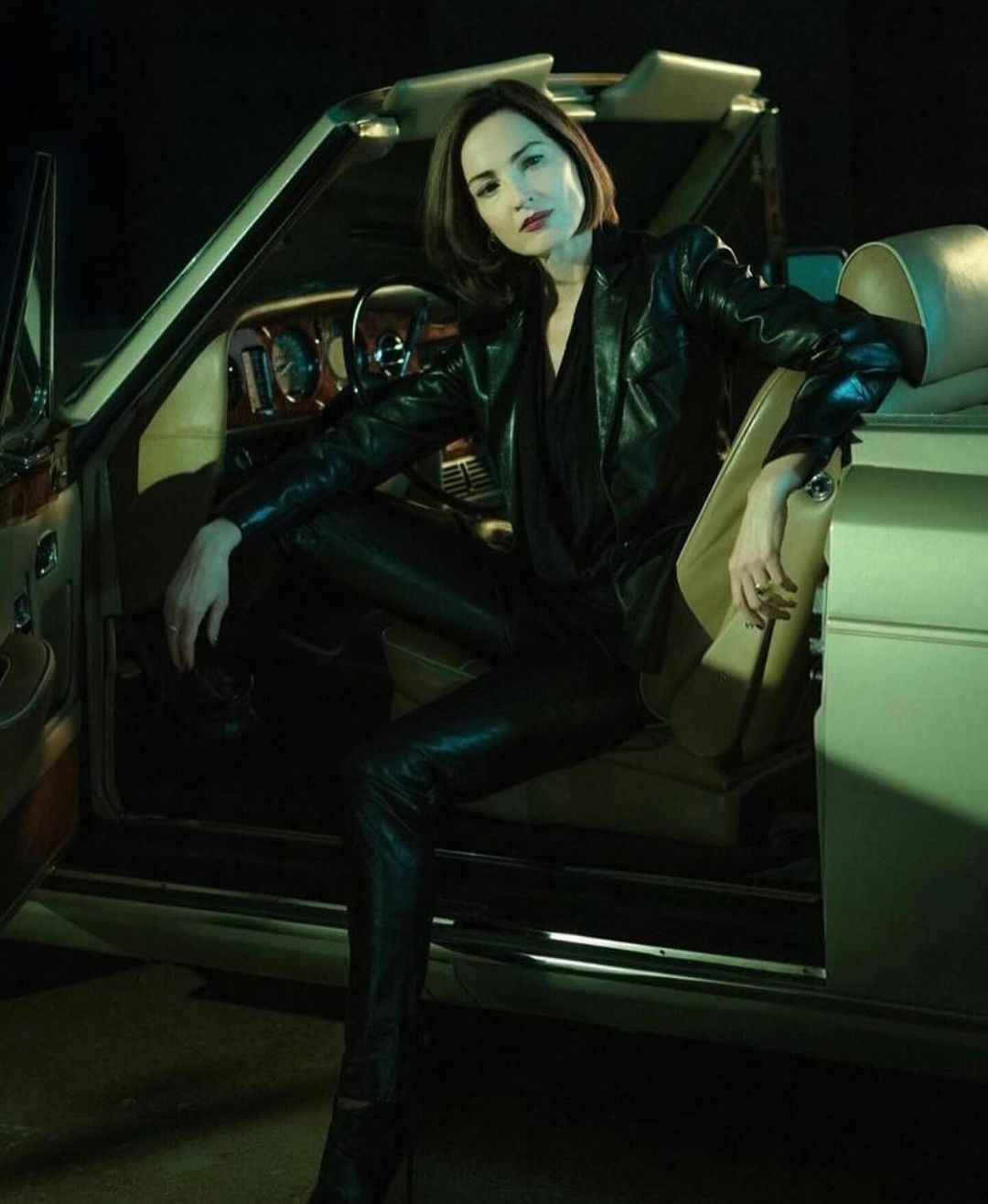 Stylish woman in a black leather jacket and pants poses inside a classic convertible car, creating a high-fashion automotive scene.