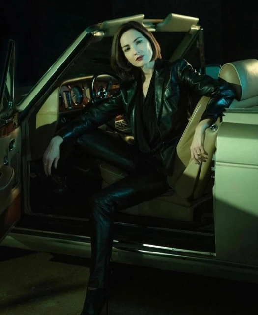 Stylish woman in a black leather jacket and pants poses inside a classic convertible car, creating a high-fashion automotive scene.