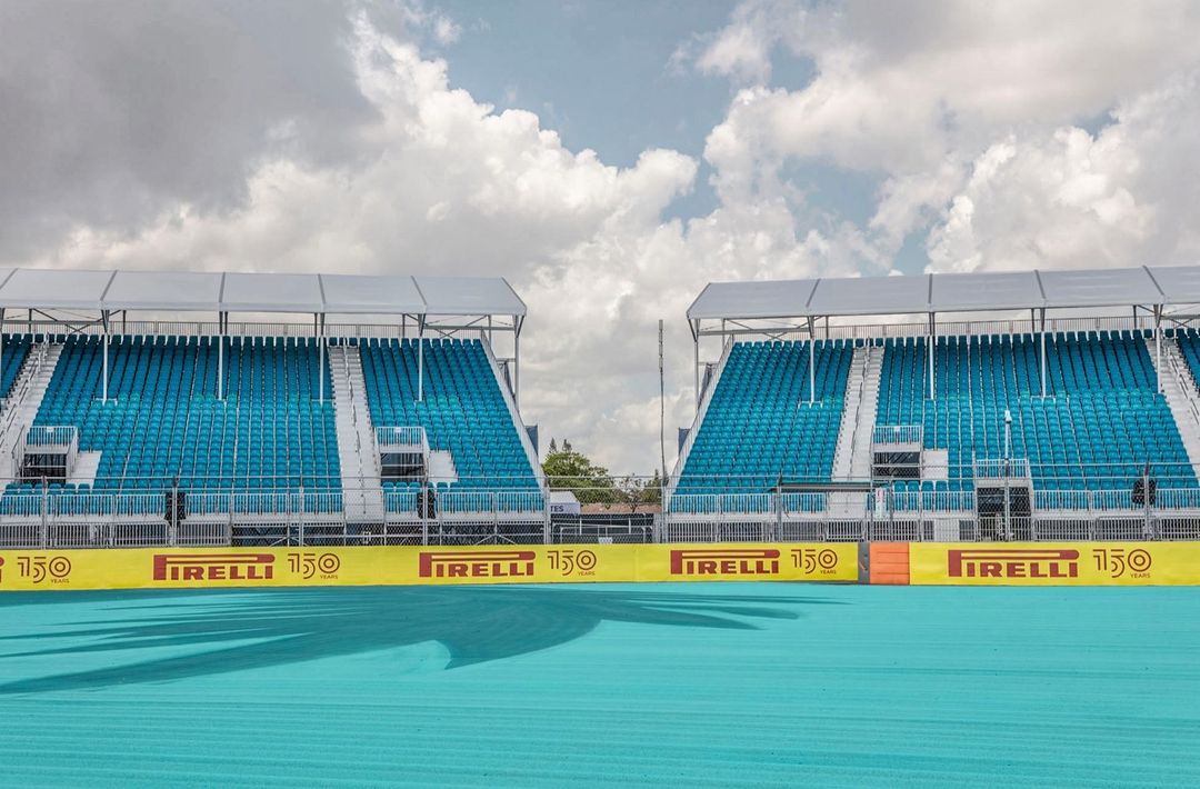 Outdoor shot of empty turquoise stadium seating under a cloudy sky. Pirelli banners line the front of the bleachers in yellow and red.