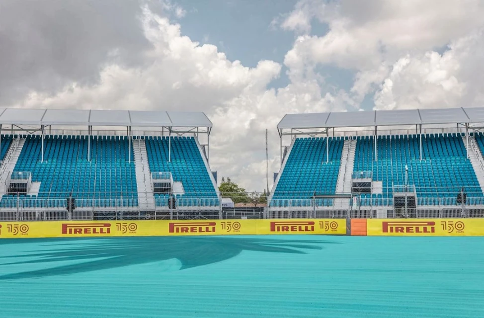 Outdoor shot of empty turquoise stadium seating under a cloudy sky. Pirelli banners line the front of the bleachers in yellow and red.