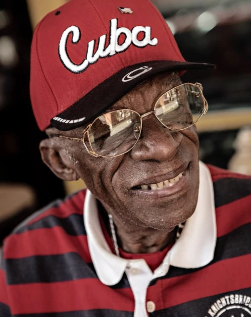 Elderly Black man in 'Cuba' baseball cap and glasses smiles warmly in a close-up portrait, showcasing details of his face and attire.