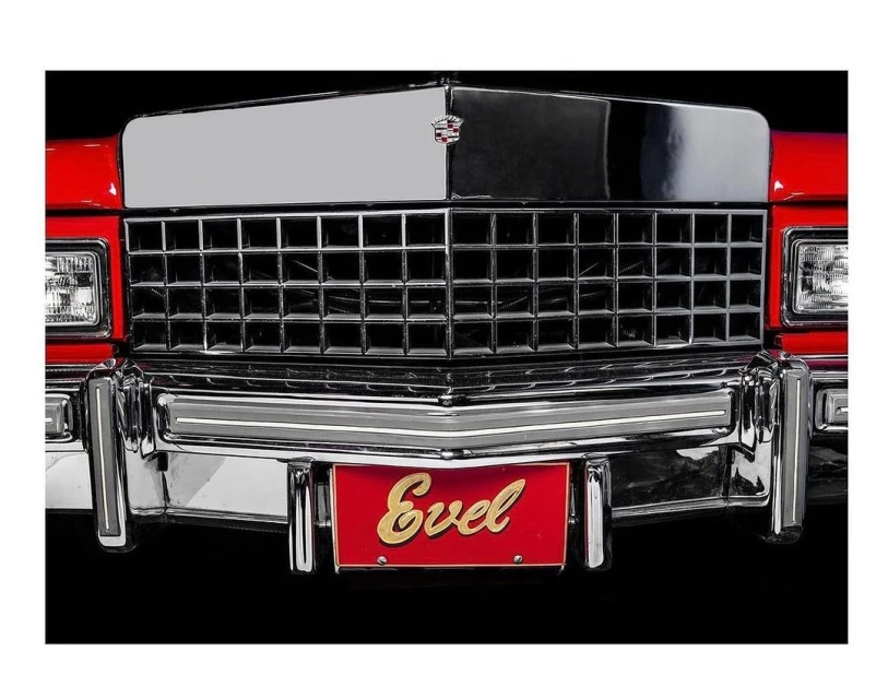 Close-up of a vintage red Cadillac with a silver grill, chrome bumper, and a red license plate with the gold text 'Evel'.