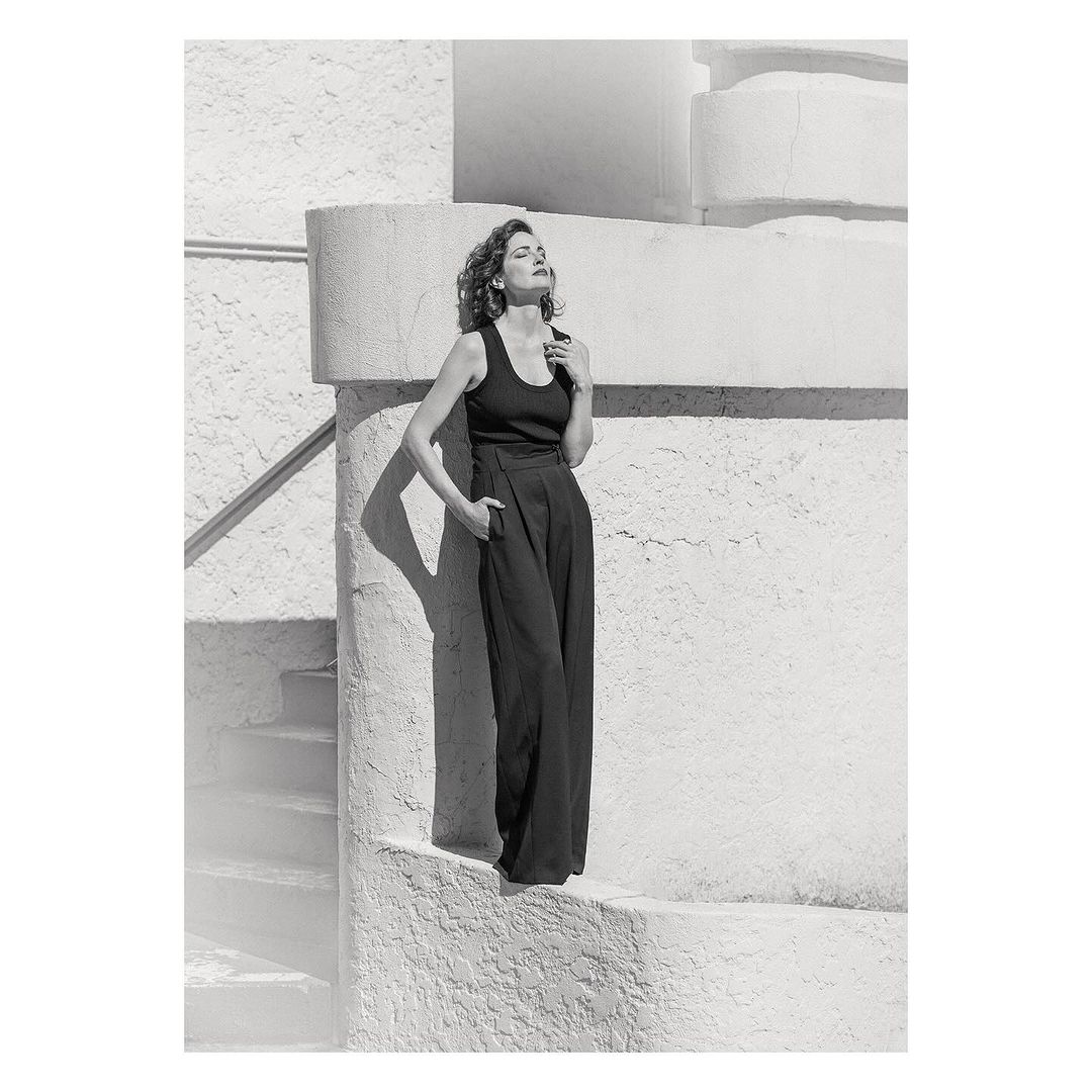 A stylish woman poses in black top and wide-leg pants, bathed in sunlight against a textured white wall with steps and railings.