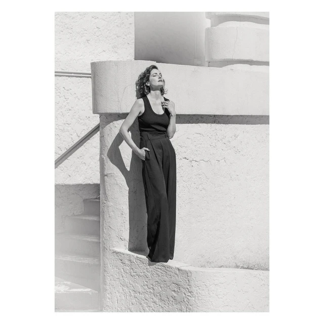 A stylish woman poses in black top and wide-leg pants, bathed in sunlight against a textured white wall with steps and railings.