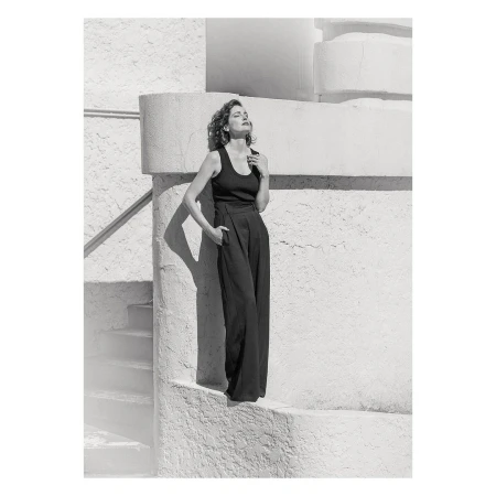 A stylish woman poses in black top and wide-leg pants, bathed in sunlight against a textured white wall with steps and railings.
