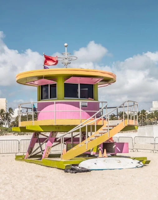 A vibrant, colorful lifeguard station on a sandy beach under a blue sky. A surfboard rests against the base of the structure.