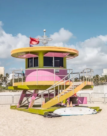 A vibrant, colorful lifeguard station on a sandy beach under a blue sky. A surfboard rests against the base of the structure.
