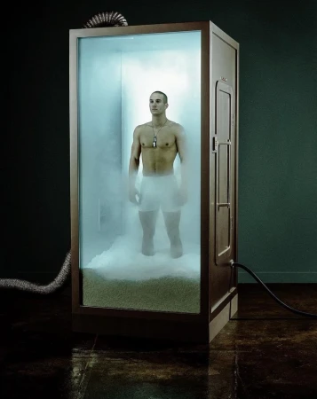 Athletic man stands in a full-body cryotherapy chamber, visible through the transparent door with vapor surrounding his lower body.