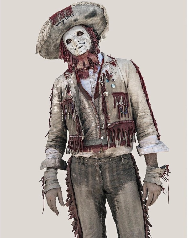 Person in a distressed costume with a mask and sombrero stands with hands cuffed in front of a neutral backdrop. 