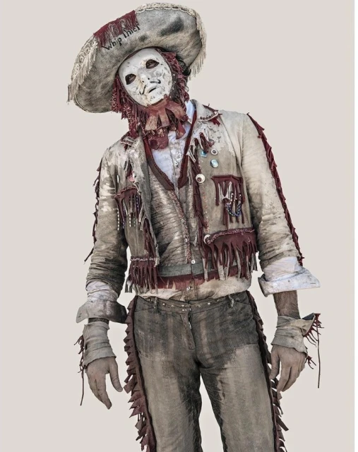 Person in a distressed costume with a mask and sombrero stands with hands cuffed in front of a neutral backdrop. 