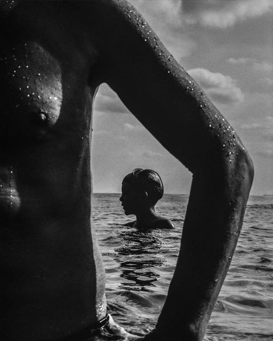 Black and white photograph framing a person in the water through the raised arm and torso of another figure in the foreground.