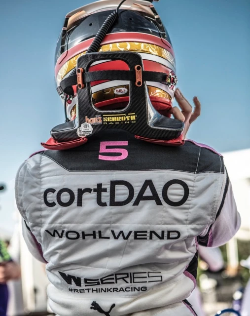 Close-up shot from behind of a racer wearing a helmet and racing suit with sponsors and text on the back, racing environment.