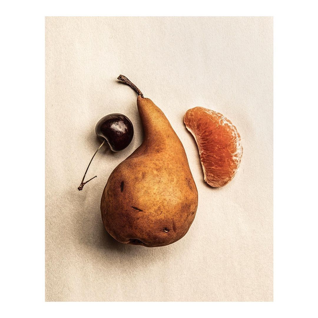 Still life shot of a pear, cherry, and tangerine segment against a light tan background in a minimalist food photography aesthetic.