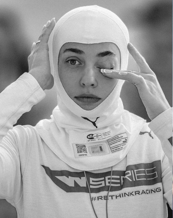A female race car driver in a white racing balaclava touches her face. Close-up shot, black and white with a blurry outdoor background.