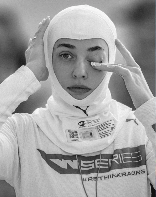A female race car driver in a white racing balaclava touches her face. Close-up shot, black and white with a blurry outdoor background.