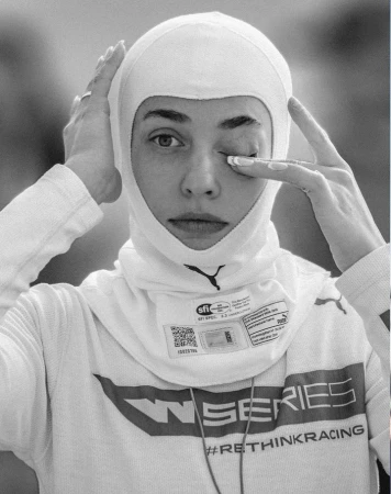 A female race car driver in a white racing balaclava touches her face. Close-up shot, black and white with a blurry outdoor background.