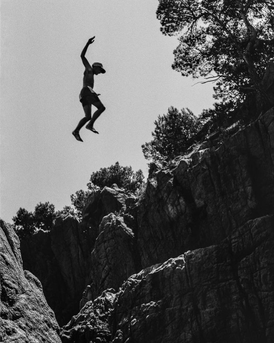 Monochrome photo of a shirtless man jumping from a rocky cliff face with trees in the background, against a clear sky.