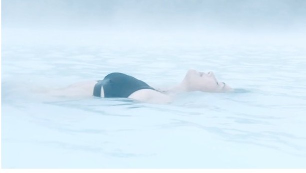 Adult woman relaxes in light blue water with a hazy, misty background, wearing a black swimsuit. Reclined back floatation.