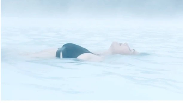 Adult woman relaxes in light blue water with a hazy, misty background, wearing a black swimsuit. Reclined back floatation.