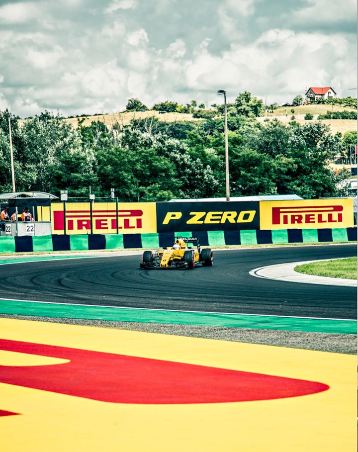 Formula 1 race car speeding along the track, surrounded by greenery and trackside signage under a cloudy sky, motorsports event.