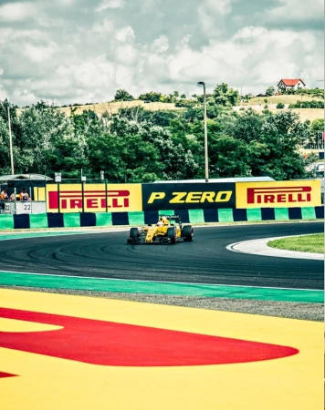 Formula 1 race car speeding along the track, surrounded by greenery and trackside signage under a cloudy sky, motorsports event.