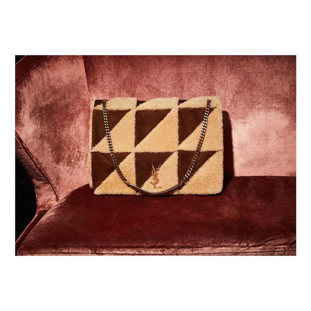 Beige and brown faux fur handbag with chain detail sitting on a burgundy velvet chair in a high-fashion, luxurious setting.