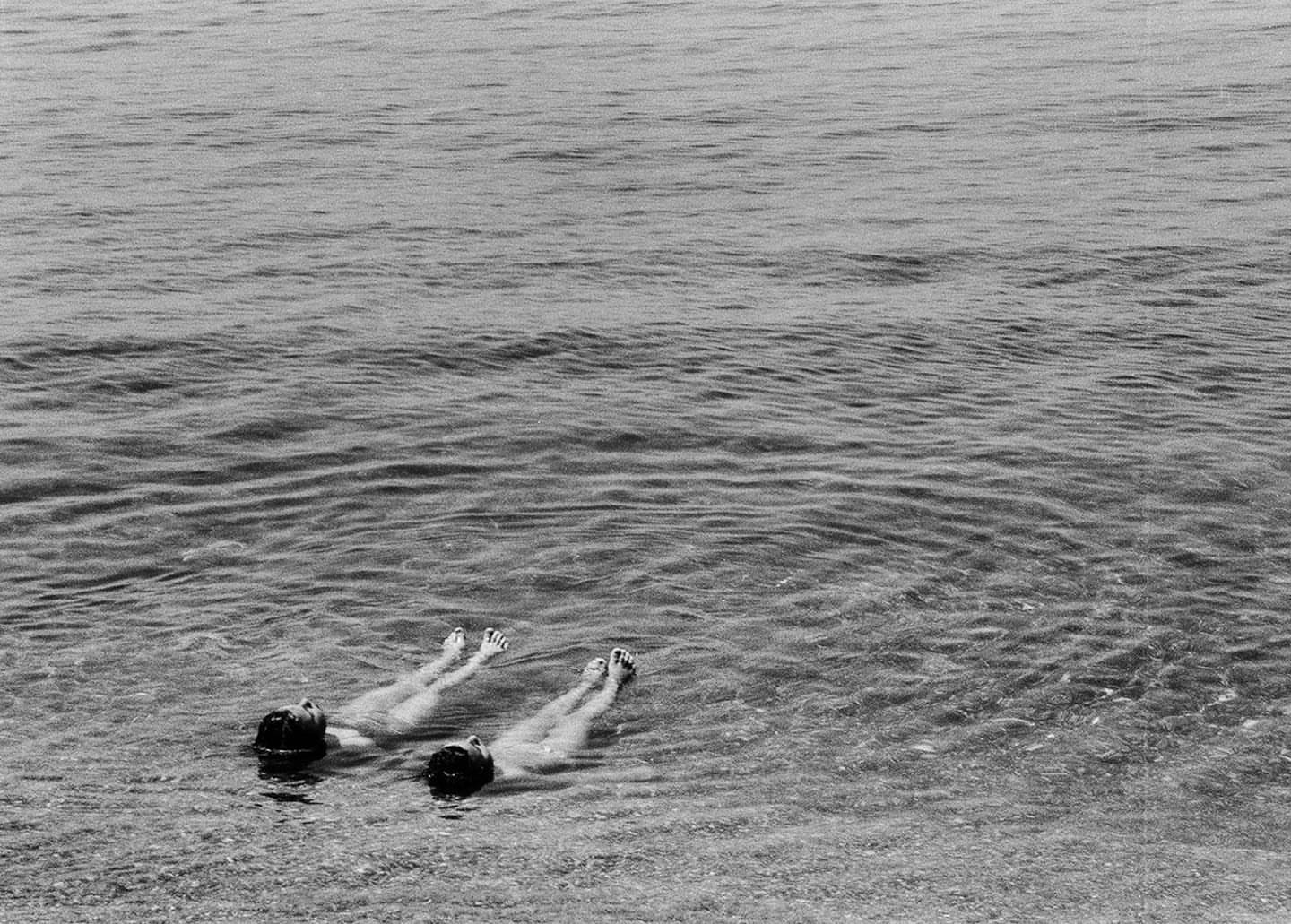 Two people floating in the ocean water, head up and facing skyward in black and white, on a cloudy day with ripples on the water's surface.
