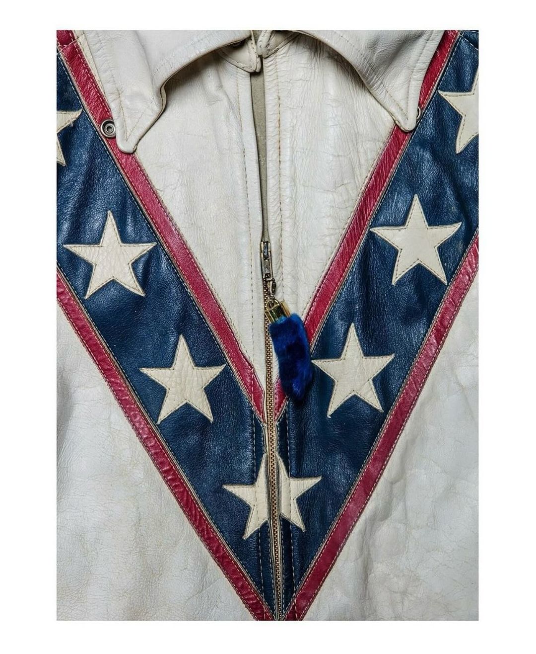 Close-up of a white leather jacket with a V-shaped blue and red pattern featuring white stars, fastened with a central zipper.